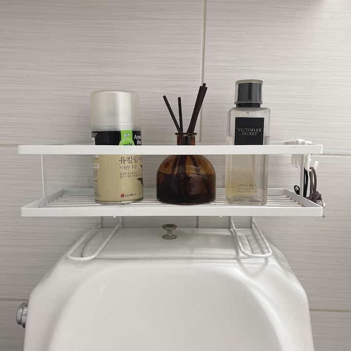 White tiled bathroom featuring white shelf over toilet tank with diffuser and fragrance items for storage