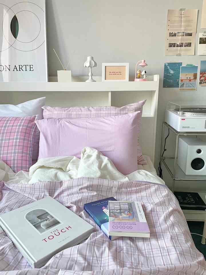 Soft pastel pink and white bedroom featuring a bed with multiple pillows and duvet, framed art and posters on the wall creating a cozy atmosphere