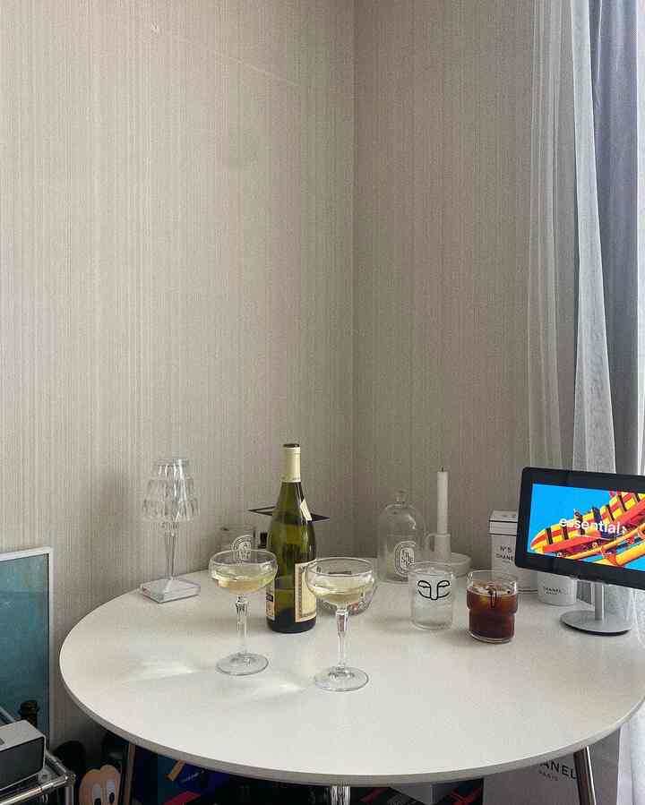 Bright-toned small room with a white round dining table featuring wine glasses, a wine bottle, candles, and transparent lighting, creating a cozy home bar