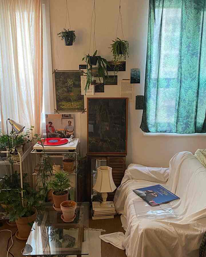 ナチュラルな色調のワンルームリビング、白いソファとガラステーブル、多数の植物とヴィンテージの小物が配置された落ち着いた空間