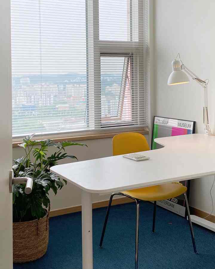 A compact home office with white tones and a yellow accent chair, featuring blinds on the window and a large plant for a cozy atmosphere