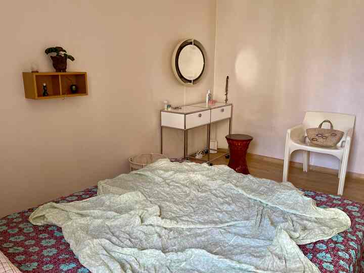 Simple bedroom with beige walls and wooden floor, featuring a white vanity with round mirror and a bed with spread duvet cover at center