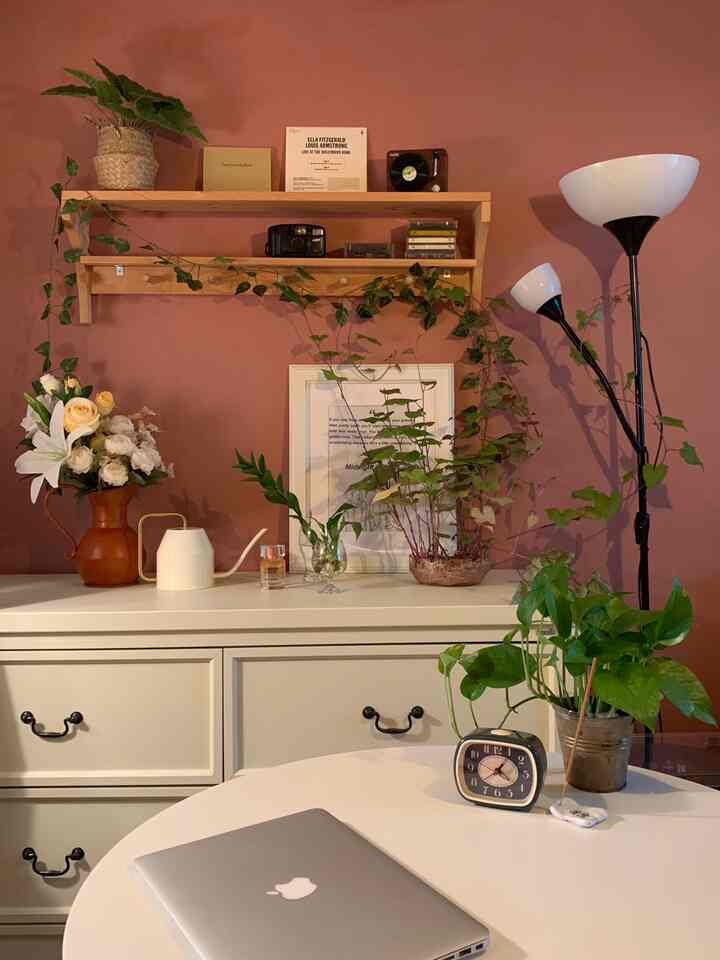 A cozy living room with brown walls, white dresser and round table, various plants, and a floor lamp creating a natural atmosphere
