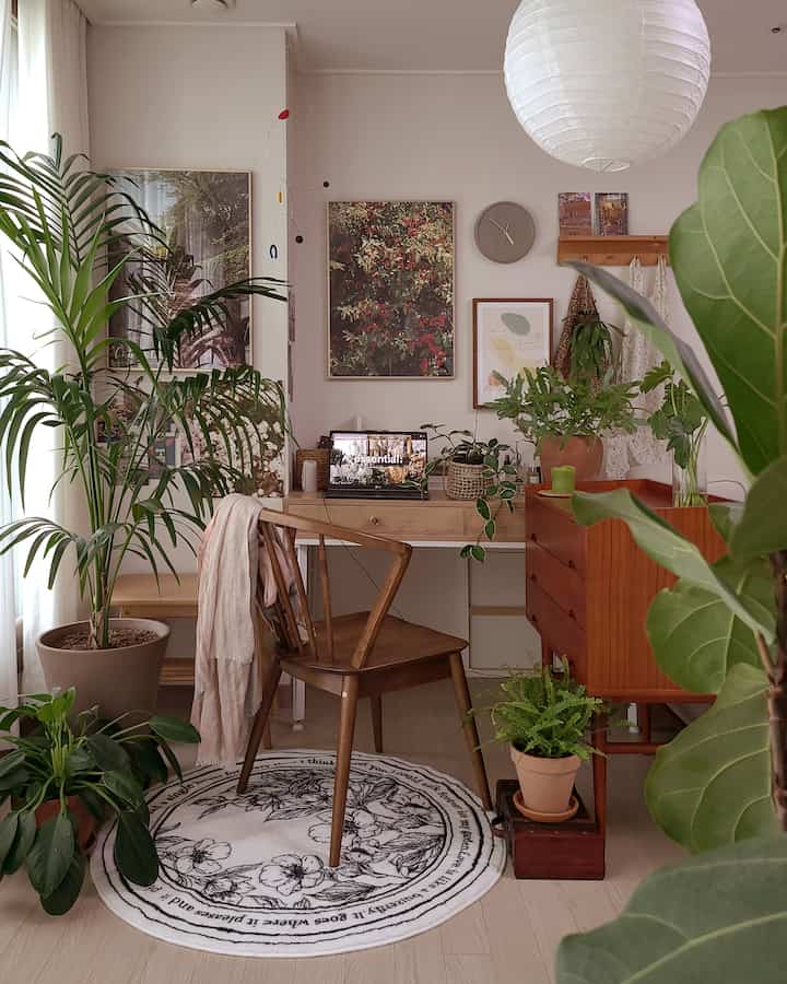 Natural toned living room featuring wooden desk and chair, surrounded by various plants creating a cozy and healing atmosphere