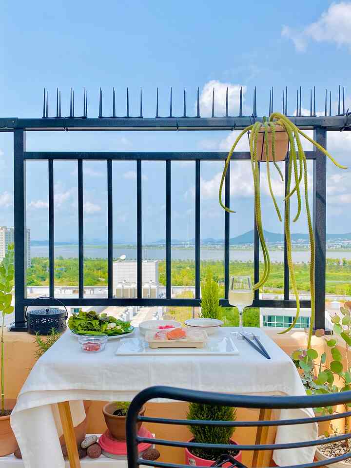 青空と緑の景色が広がるバルコニー、白いテーブルクロスが掛かったサイドテーブルと吊るされたサボテンが調和する居心地の良い一人用の食事空間