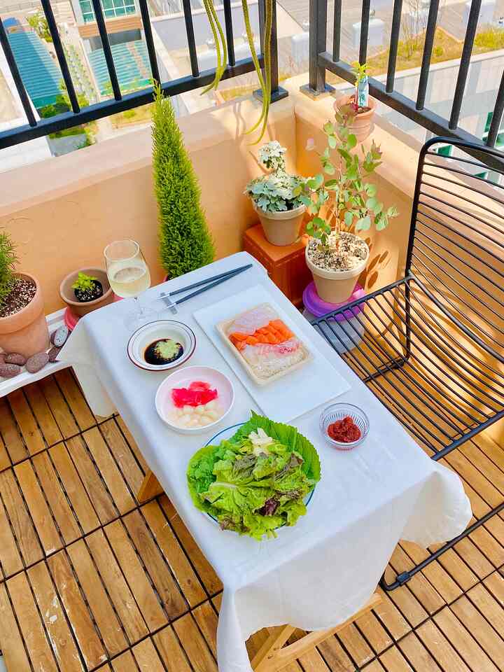 Balcony featuring a white tablecloth-covered table, wood tone deck flooring, and multiple plants with a meal set up