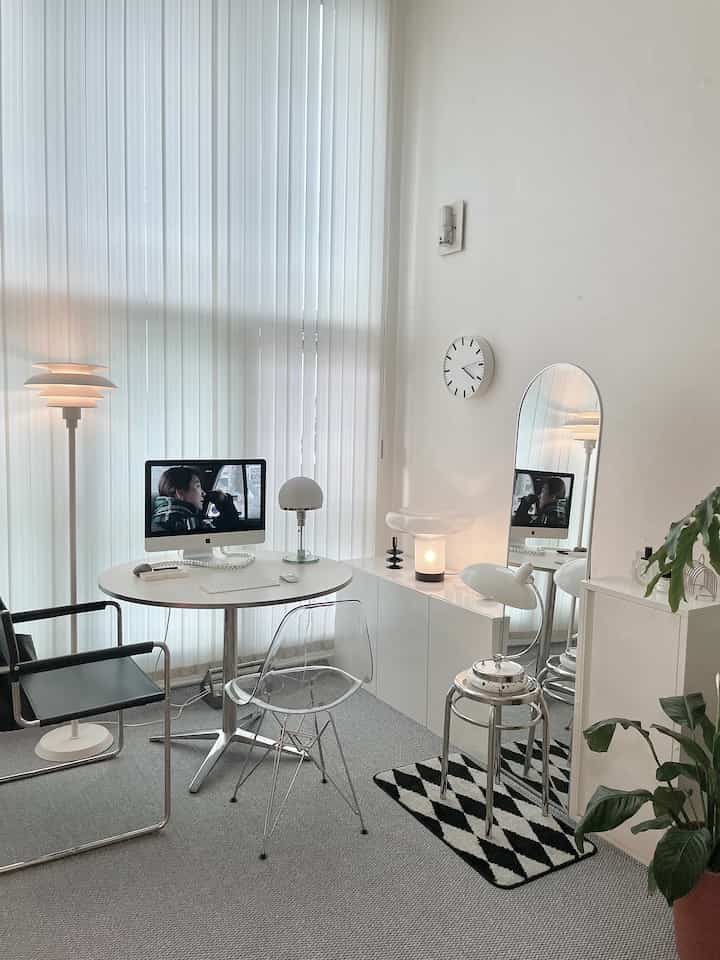 White-toned studio apartment home office featuring a round desk with iMac, transparent chair, and modern lighting elements