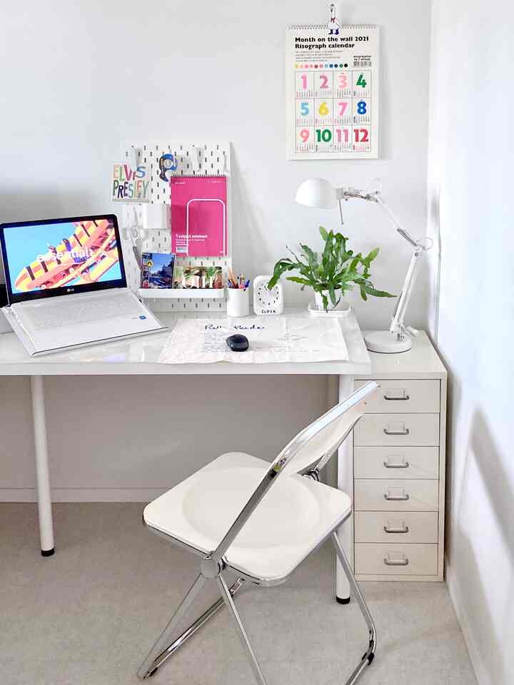 White-toned home office space featuring a desk, office chair, drawer cabinet, pegboard, and green plant creating a clean workspace
