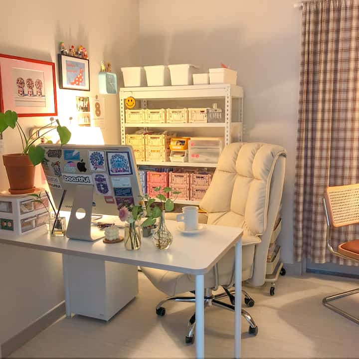 White and beige toned workspace featuring an iMac computer, office chair, metal shelving with color-coded storage boxes in a tidy cozy room
