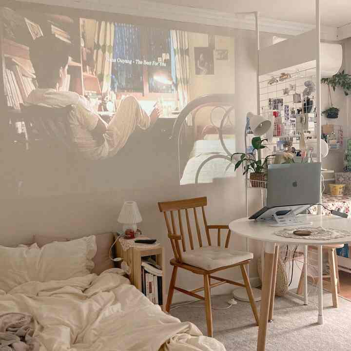 A neutral wood-tone minimal studio apartment featuring a wooden chair, round table, and a projector screen displaying an image on the wall