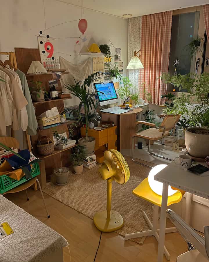A small room with white walls and wood tone furniture, featuring a desk with a yellow fan in the center, various plants, and pink curtains creating a cozy atmosphere