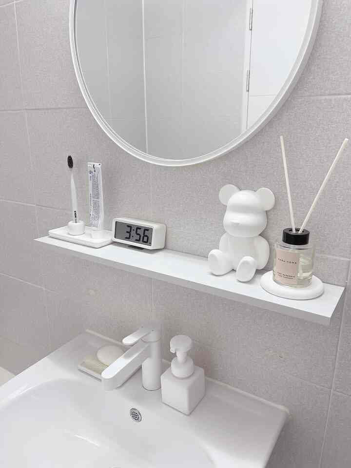 White and light gray tone bathroom featuring a round mirror, white sink with diffuser and digital clock arranged in a simple modern interior