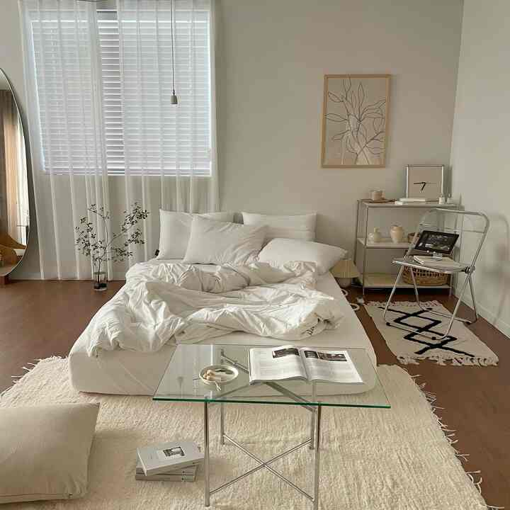 白とクリーム色調のモダンな寝室、中央にベッドとラグ、ガラステーブルと透明な椅子が配置された印象的な空間