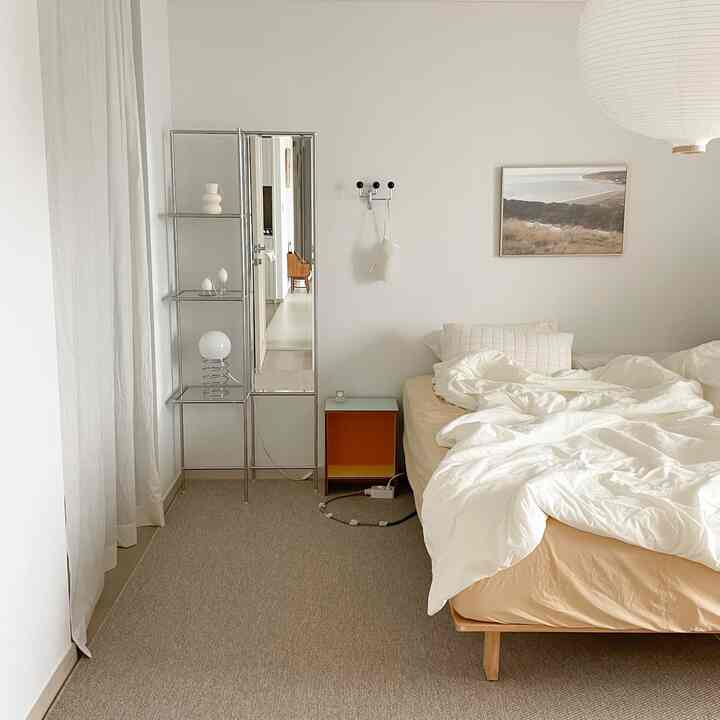 白とベージュのトーンの寝室で、白いカーテンとベッド、壁にポスターと金属製の行動ラックと全身鏡が配置された自然な雰囲気のインテリア