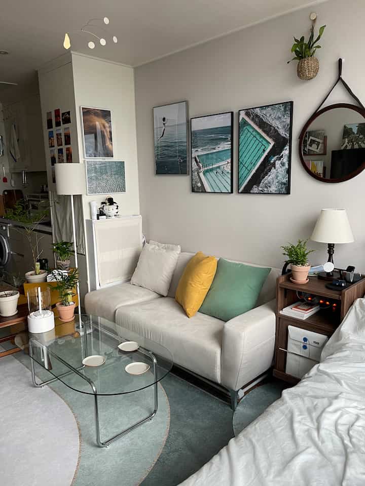 白と緑のトーンのワンルームリビング兼寝室、ソファやガラステーブル、クッション、壁のアートが配置された雰囲気ある空間