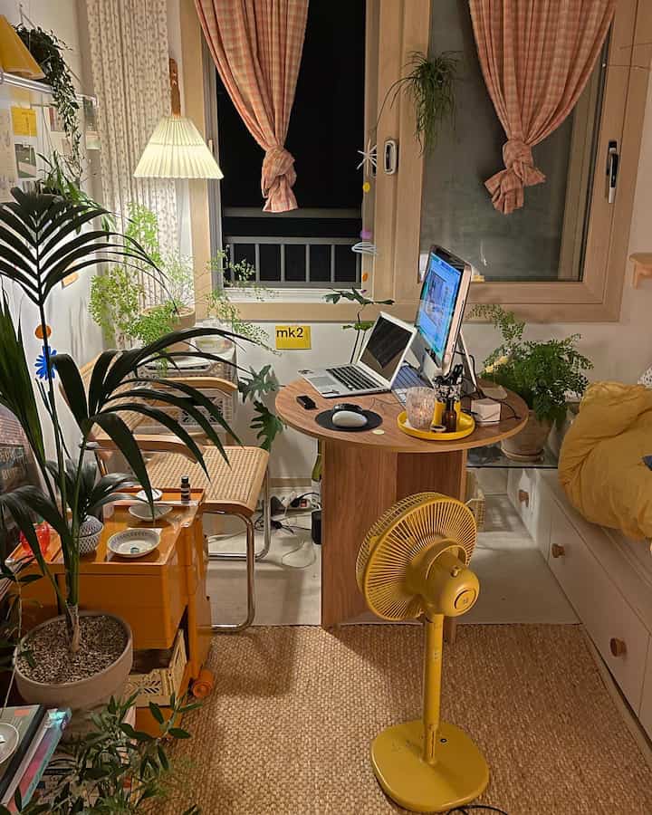 Natural toned small bedroom home office featuring a round wooden table, rattan armchair, and numerous plants creating a cozy atmosphere