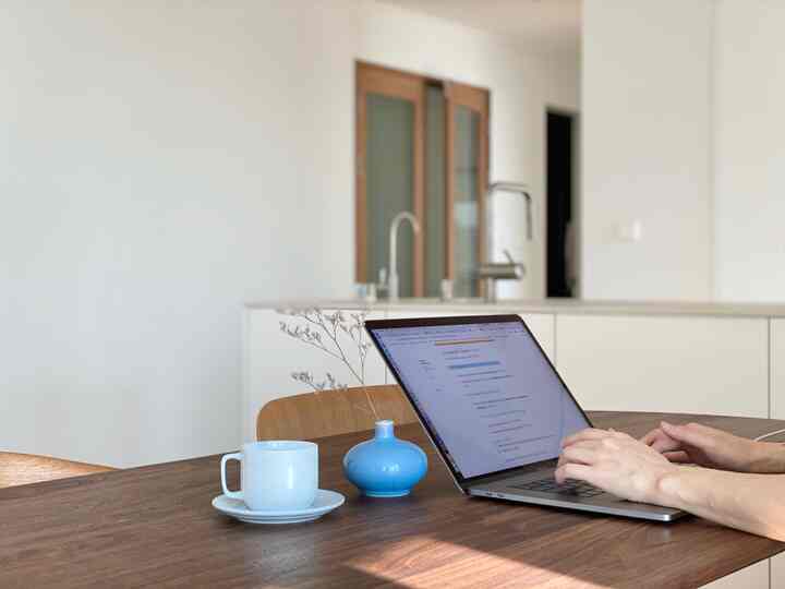 Modern natural studio apartment with white and brown kitchen and dining table, person working remotely on MacBook