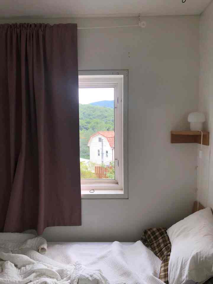 Soft white and brown toned bedroom in a small studio apartment, featuring brown blackout curtains beside the window and a wall-mounted corner shelf with a mushroom-shaped mood lamp