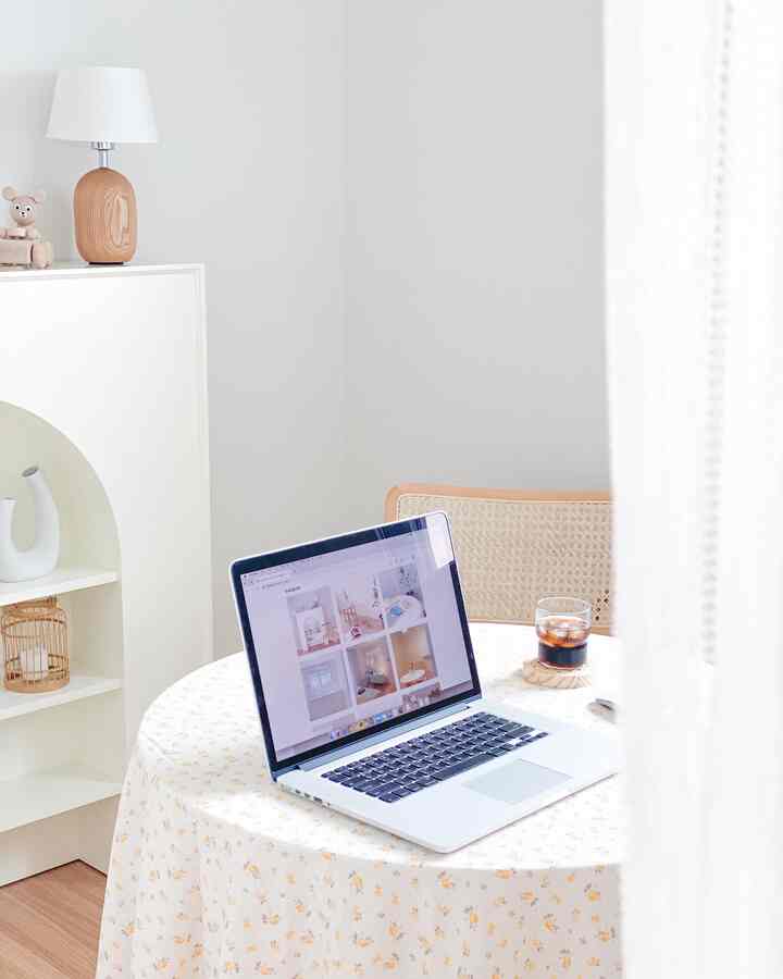 Small living space in white and brown tones featuring a floral tablecloth with laptop and iced coffee, rattan armchair, and shelf creating a cozy atmosphere