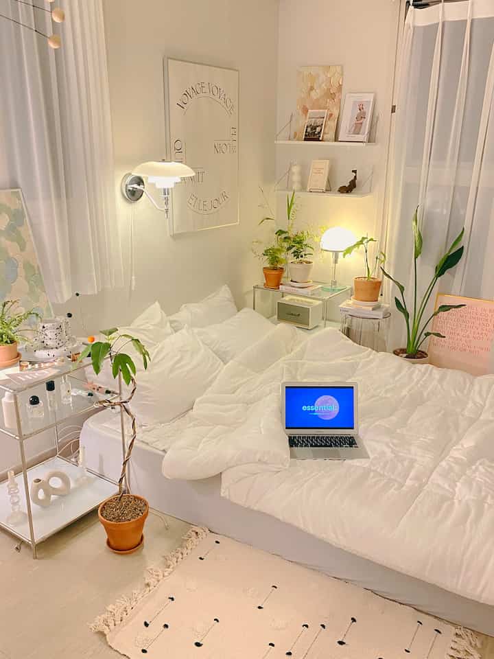 White-toned studio bedroom featuring a cozy bed with duvet cover, potted plants, and minimalist decor