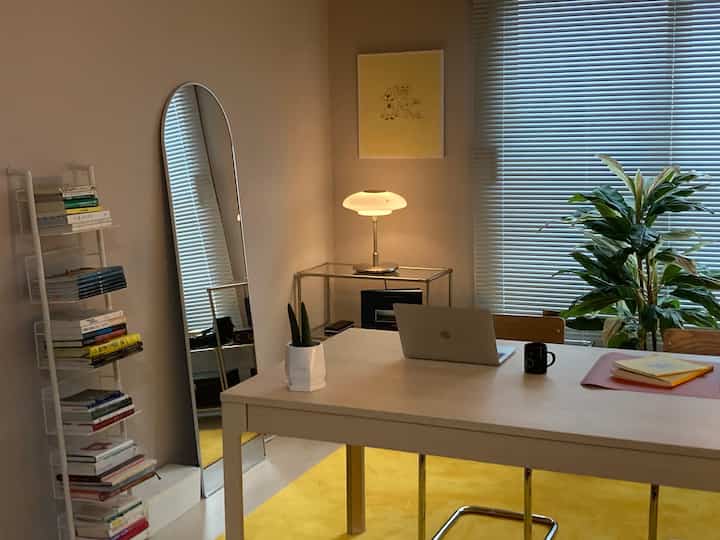 Bright yellow rug and white furniture, bookshelf, tall mirror, and plant arranged in cozy home office space