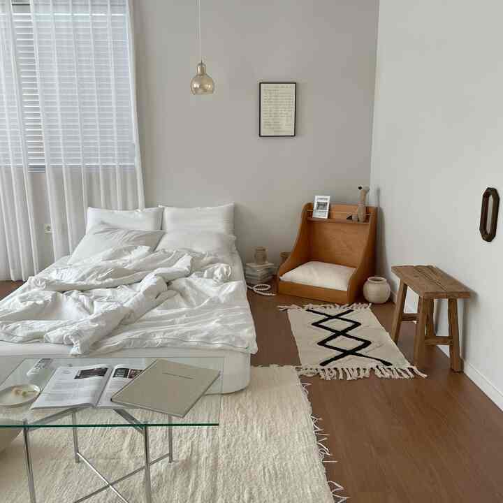 A white and wood-toned bedroom featuring white bedding, glass table, wooden stool, and rugs in a simple modern style