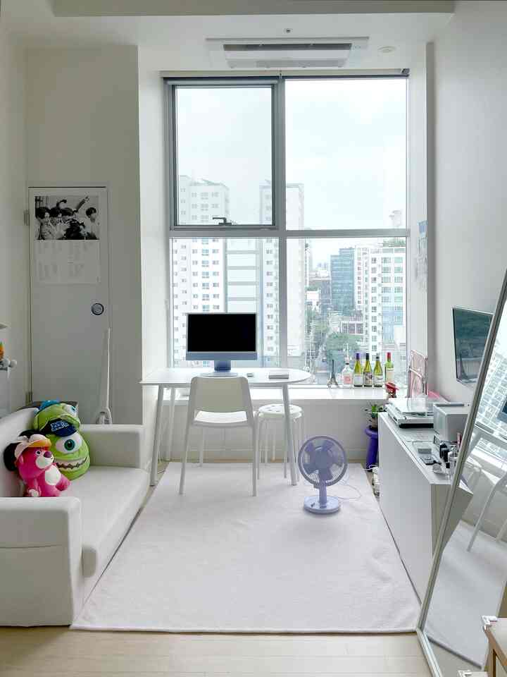 Bright white-toned studio apartment home office space featuring desk and chair by a large window, accompanied by sofa and rug creating a cozy atmosphere