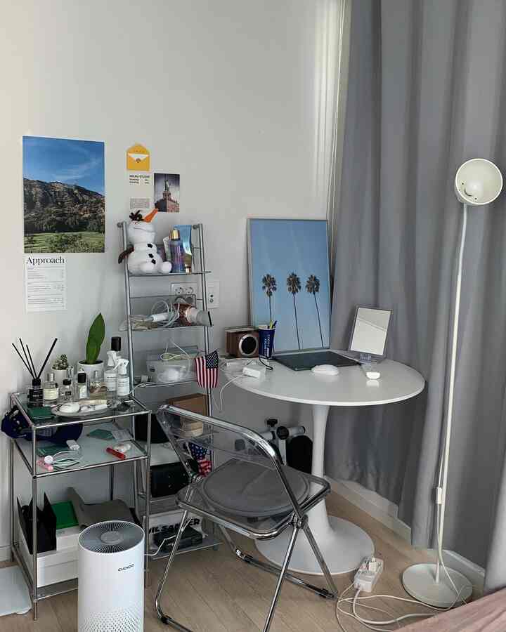 Compact room with gray curtains and white walls, featuring a round table, transparent chair, and metal shelving for a practical setup