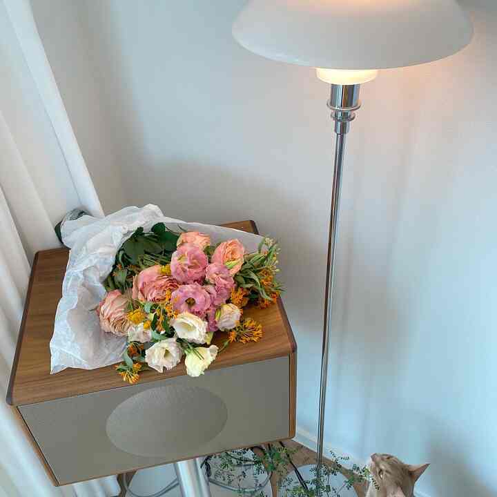 Cozy plant interior space with wood-toned speaker topped by a bouquet of flowers and elegant floor lamp against white walls and curtains