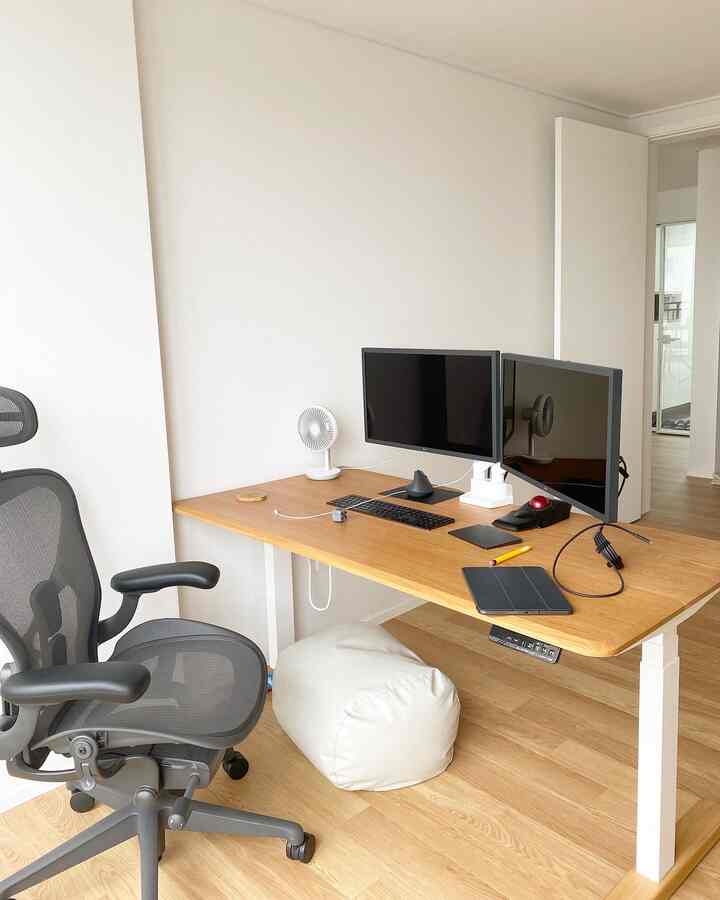 明るい木目調と白を基調とした書斎、モーションデスクとデュアルモニター、エアロンチェアが配置された清潔感のあるホームオフィス