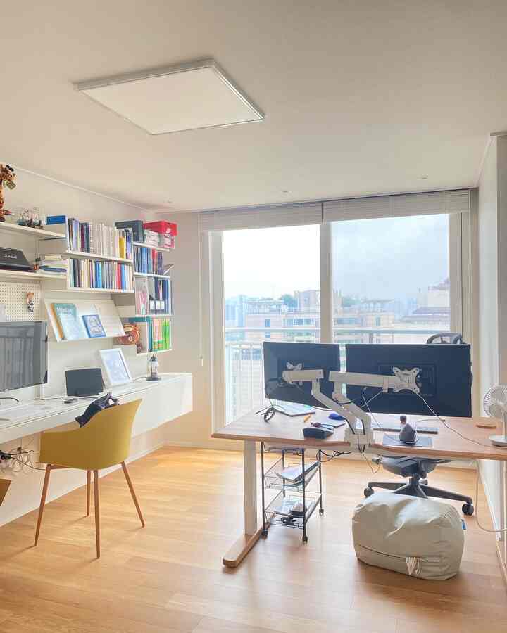 A bright and tidy home office with white walls and wood-tone floor, featuring dual monitor desks and a bookshelf