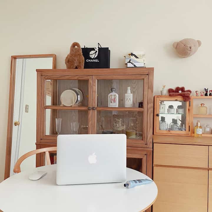 White and natural wood tone bedroom space featuring wooden cabinets and glass display cabinet with a round table and MacBook, cozy home office atmosphere