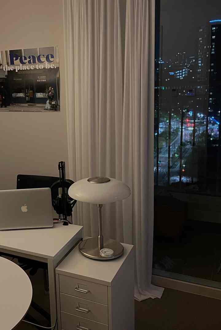 White-toned compact home office with MacBook, table lamp, blackout curtains, and poster creating a quiet nighttime ambiance