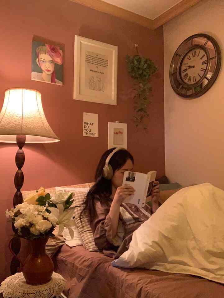 暖かいナチュラルカラーの壁とヴィンテージ照明がある寝室で、ベッドに寝そべって本を読む姿が印象的な空間