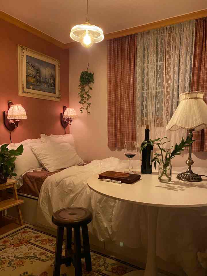 A pink-toned studio bedroom featuring white bedding, vintage decor, and warm lighting creating a cozy hotel-like atmosphere