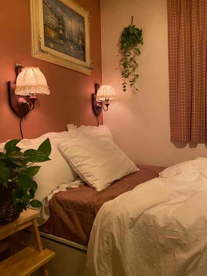 A bedroom with pink walls and wood-tone stool, featuring white bedding and wall sconces creating a cozy atmosphere