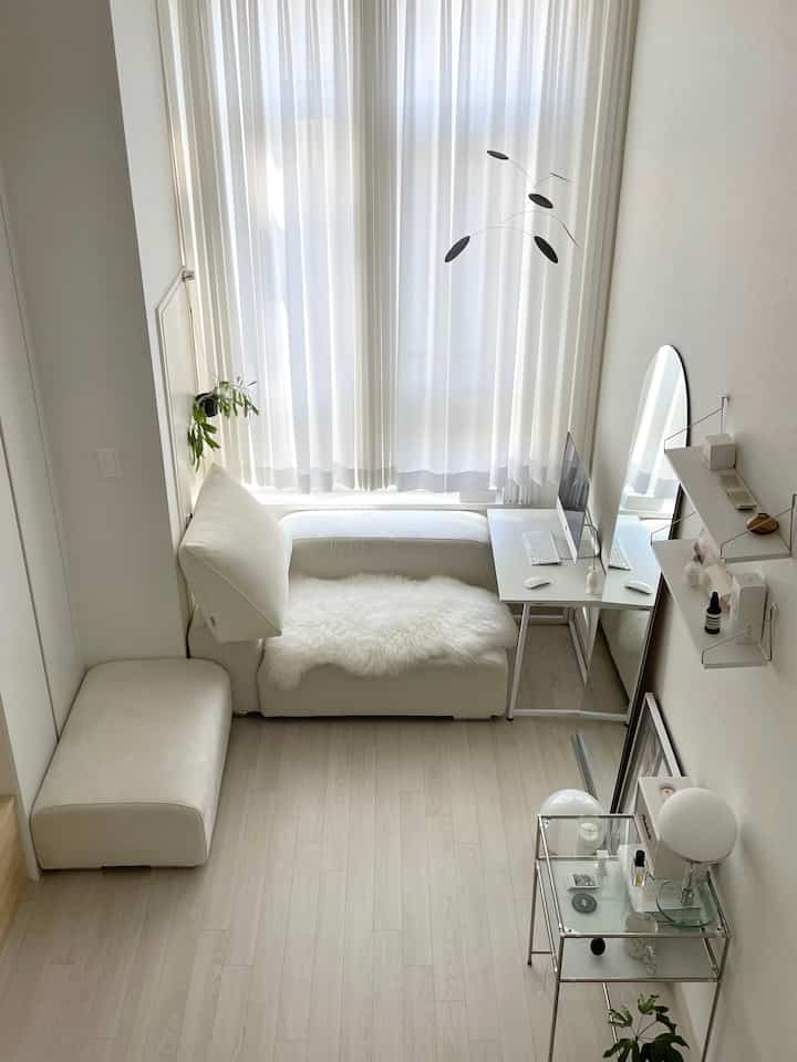Bright white-toned living room featuring modular sofa center-left, sheer curtains on window, small work table and full-length mirror on right, with minimal décor