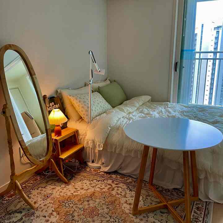 A cozy single household bedroom featuring white bedding, wood tone full-length mirror, and round table, blending light and natural tones