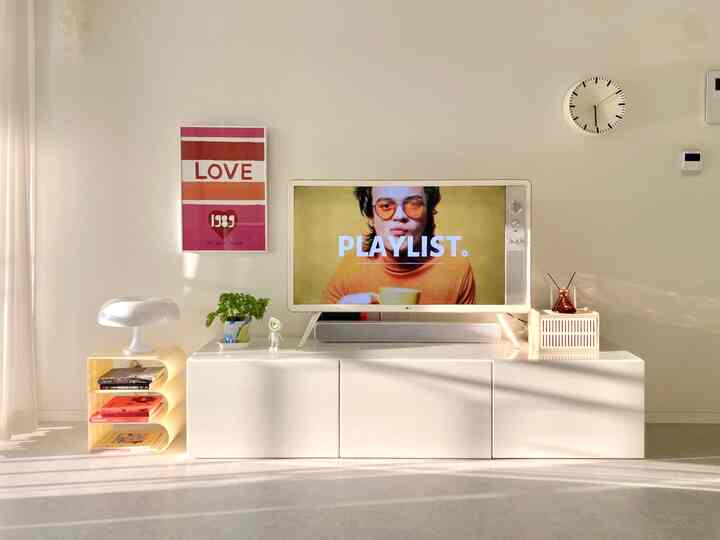 Bright white-toned living room featuring a central white TV stand with a clock on the right wall and a yellow bookshelf and table lamp on the left, creating a clean atmosphere