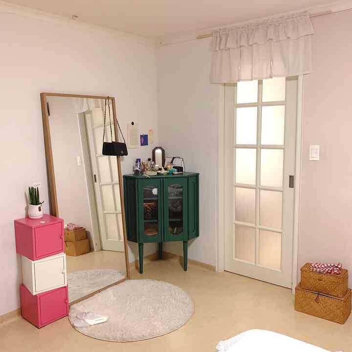 白い壁と自然なトーンの床の寝室、角に置かれた緑のコンソールと大きな丸いラグ、木枠の大きな鏡が配置された雰囲気のあるインテリア