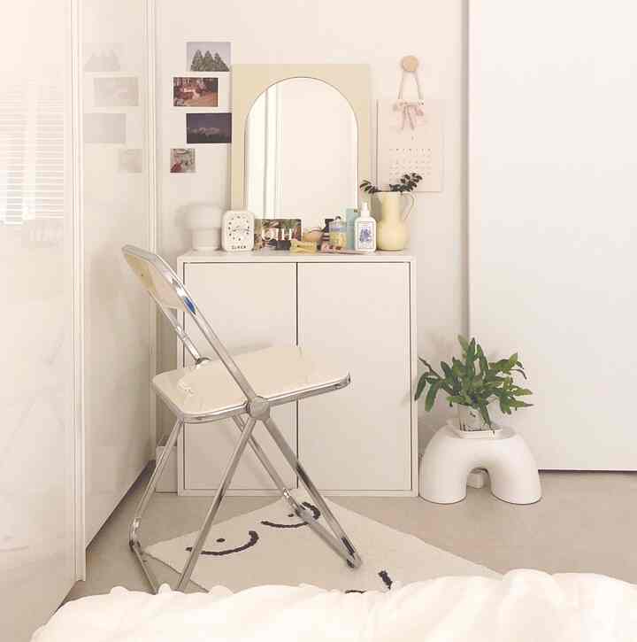 White-toned bedroom corner featuring a vanity with an arched mirror and folding chair in a clean, minimal space