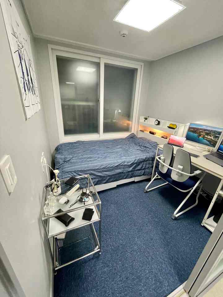 白と青のトーンが特徴の小さなワンルーム寝室、机とオフィスチェアが配置されたミニマルで効率的な空間