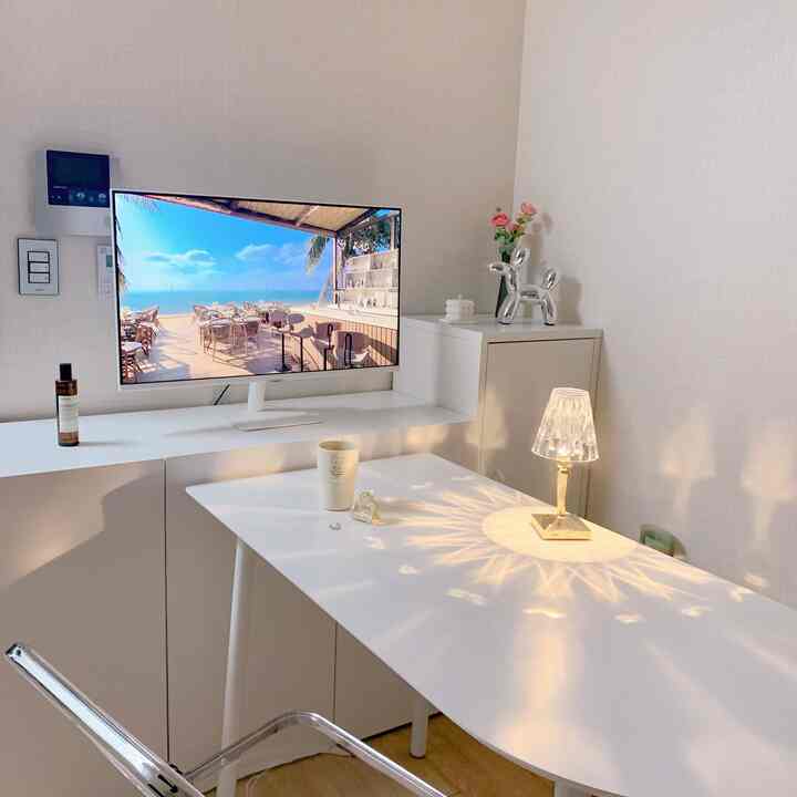 White-toned home office space featuring a large desk with monitor stand, transparent chair, and decorative table lamp creating stylish ambiance