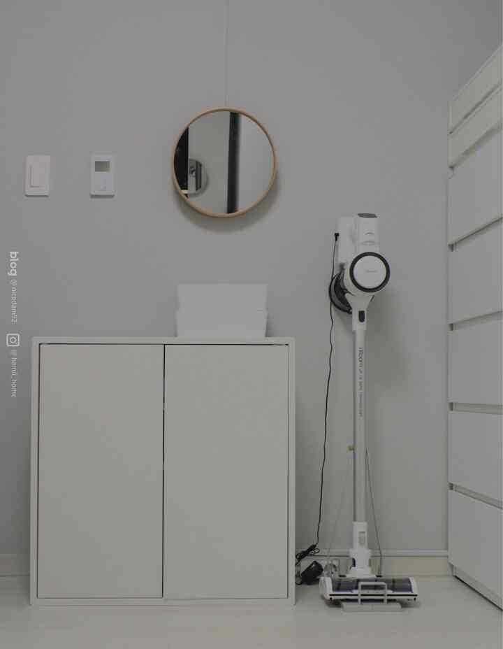 White-toned walk-in closet featuring a round wall mirror, tidy dresser, and wall-mounted cordless vacuum cleaner with a simple organized look