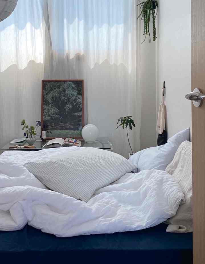 柔らかいホワイトトーンの寝室、白い寝具と枕、透明なテーブルの上に額縁と植物が置かれた落ち着いた空間