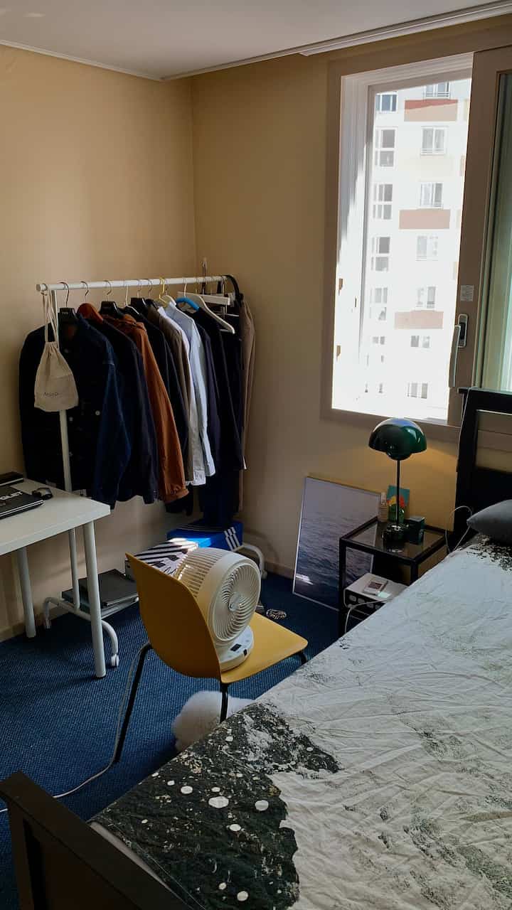 Modern studio bedroom with navy rug and yellow chair, featuring clothing rack and nightstand