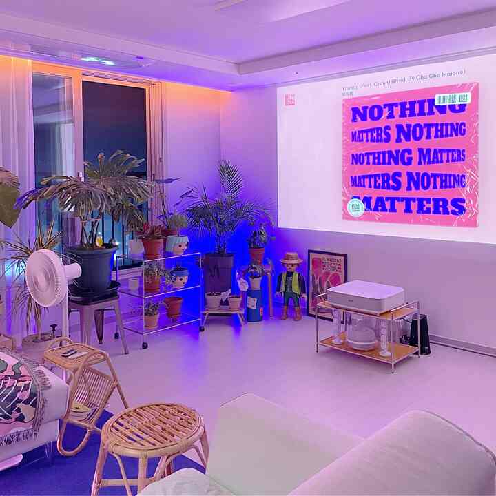 Purple-lit living room with white flooring, rattan stools, various plants, and a home cinema setup featuring a projector screen