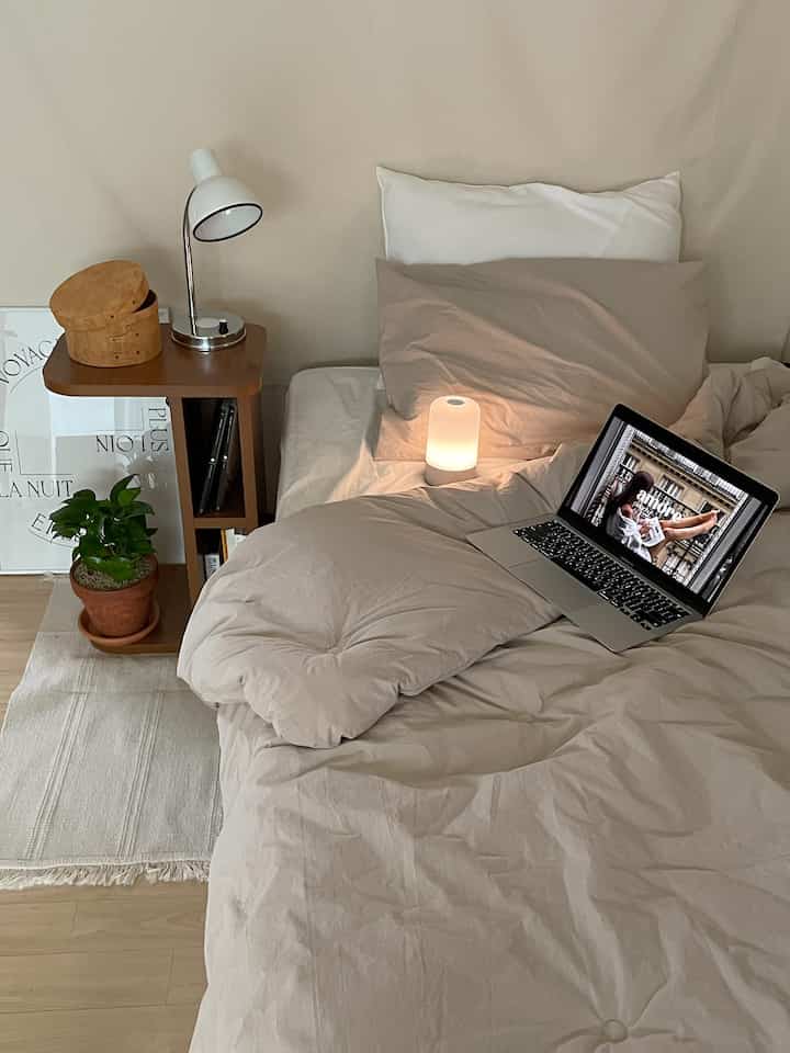 Cozy beige-tone bedroom featuring a comfortable bed, wooden nightstand, soft lighting, and a green plant creating a natural atmosphere