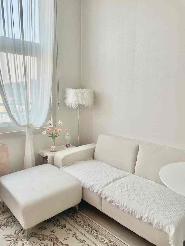 白を基調としたワンルームのリビング、ソファとスツール、リネンカーテン、羽のランプと植物が配置された落ち着いた印象的な空間