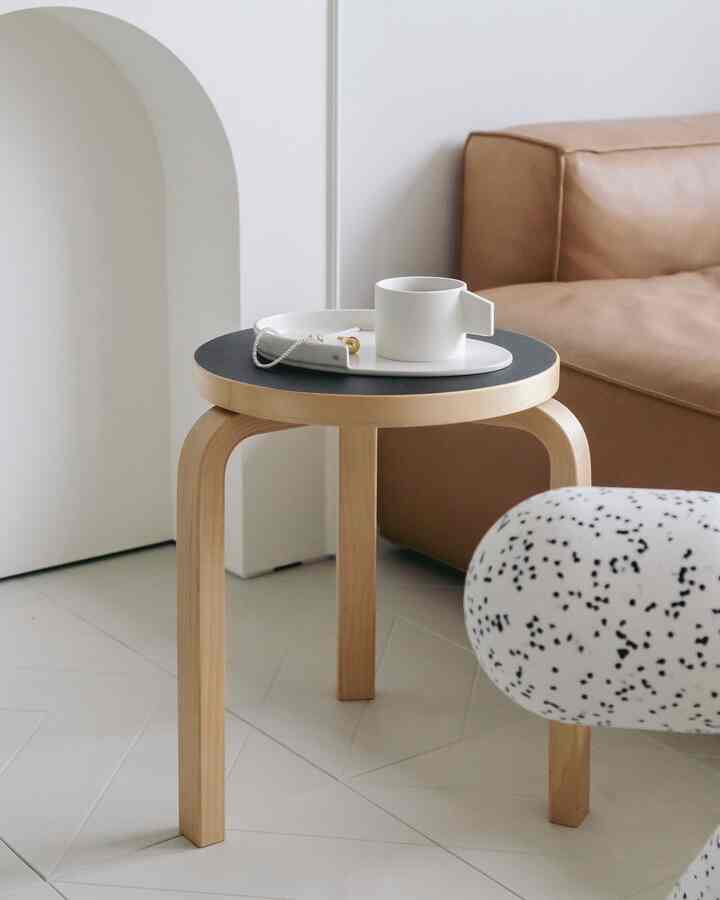 ホワイトの壁と床、ブラウンのレザーソファ、木製のスツールとトレイが配置されたシンプルでモダンなリビングルーム